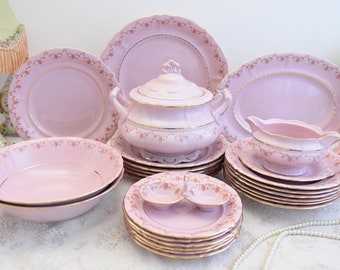 Op de afbeelding: Een roze en gouden porseleinen servies met een bloemenpatroon. De set bevat borden, kommen, een serveerschaal, een sauskom en een suikerpot.