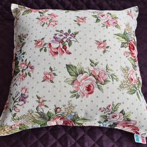 May include: A white pillow with a floral pattern featuring pink and red roses, green leaves, and small white polka dots. The pillow has a white border and is made of a soft, textured fabric.