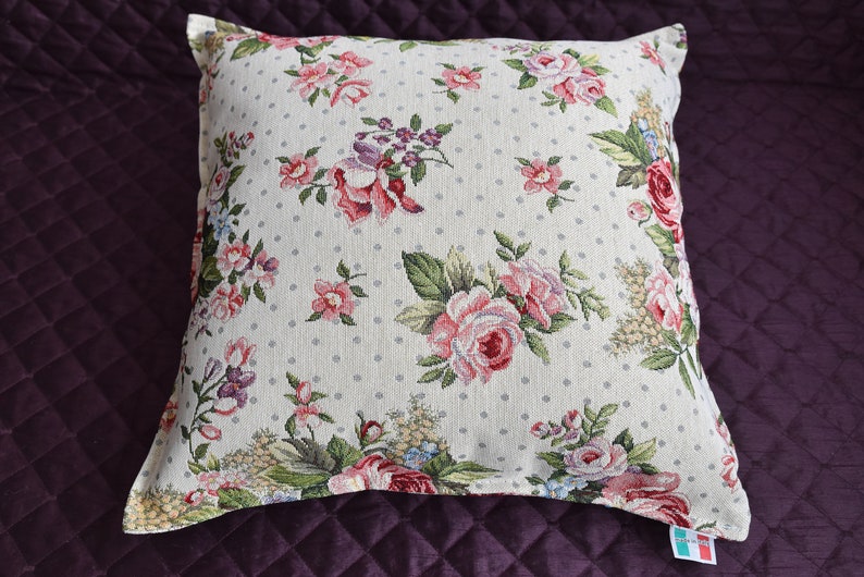 May include: A white pillow with a floral pattern featuring pink and red roses, green leaves, and small white polka dots. The pillow has a white border and is made of a soft, textured fabric.