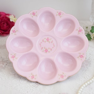 May include: A pink ceramic egg plate with a floral design. The plate has six individual compartments for holding eggs.