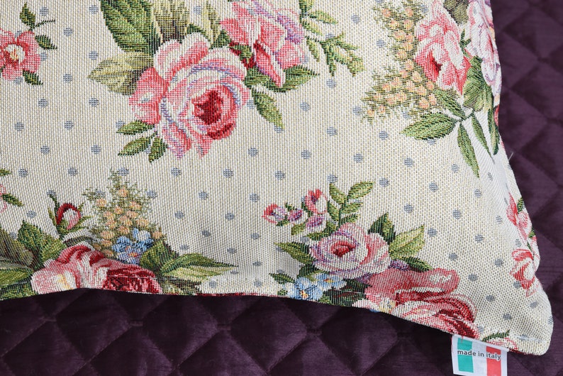 May include: A cream-colored throw pillow with a floral pattern featuring pink roses, green leaves, and blue accents. The pillow has a white polka dot background and a "Made in Italy" label.