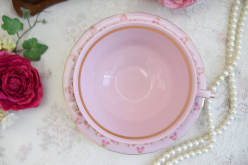 Puede incluir: Un juego de taza y platillo de t&eacute; rosa con un patr&oacute;n floral y ribete dorado. La taza de t&eacute; tiene un taz&oacute;n ancho y poco profundo y un asa. El platillo tiene un patr&oacute;n floral a juego y ribete dorado.