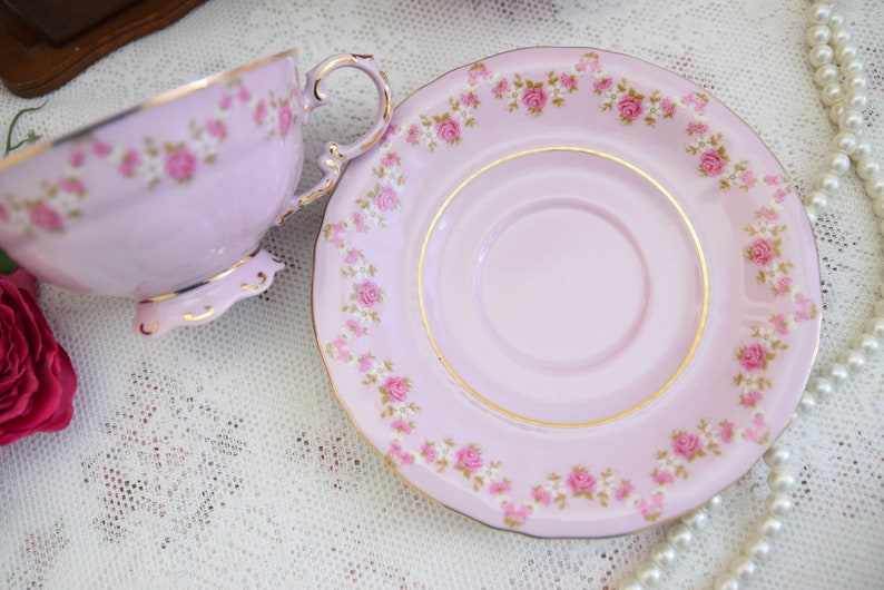 Puede incluir: Un juego de taza y platillo de t&eacute; rosa con un delicado patr&oacute;n floral. El juego presenta un borde dorado y rosas rosas con acentos blancos.