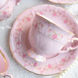 Puede incluir: Un juego de taza y platillo de t&eacute; rosa con un patr&oacute;n floral. La taza de t&eacute; tiene un borde dorado y el platillo tiene un borde festoneado.