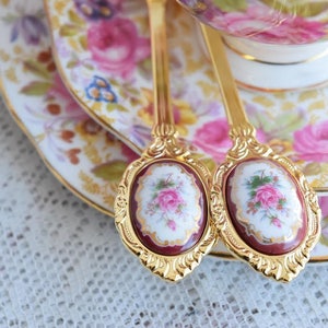 May include: Two gold-tone teaspoons with floral designs on the handles. The spoons are resting on a floral patterned saucer.