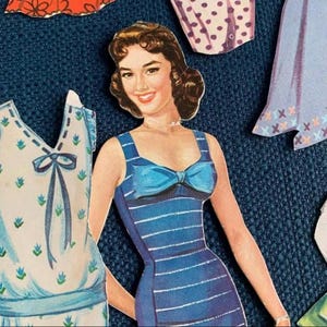 May include: Vintage paper doll with a smiling woman in a blue striped dress with a bow. Other paper doll clothing pieces are visible, including a polka dot dress and a dress with an X pattern. The paper doll is a retro design.