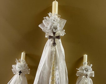 May include: Three white baptism candles decorated with lace and ribbon. The central candle features a silver cross and a larger bow.
