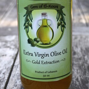 May include: A green and white label on a glass bottle of extra virgin olive oil. The label reads "Gem of El-Koura Extra Virgin Olive Oil Gold Extraction Product of Lebanon 32 oz."