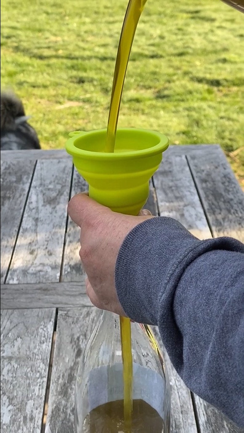 May include: A person pouring yellow liquid from a bottle into a green silicone funnel. The funnel is collapsible and has a wide opening.