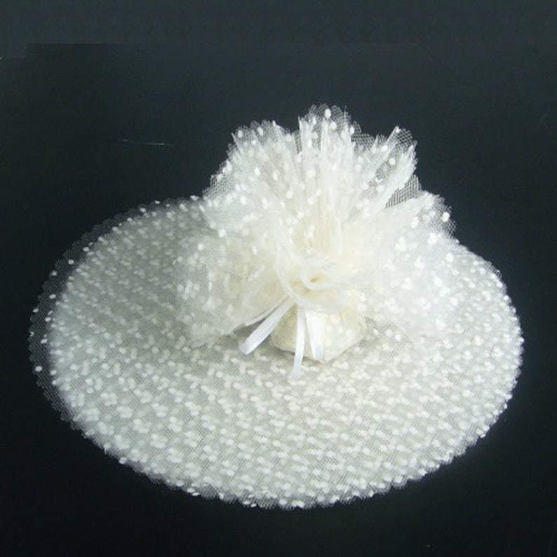 May include: White tulle fabric with small white polka dots. The tulle is gathered into a round shape with a white ribbon tied in a bow.
