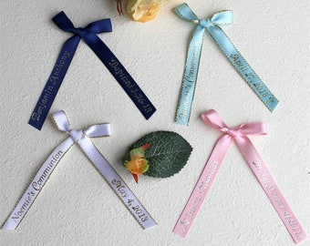 May include: Five personalised satin ribbon bows with gold lettering. The ribbons are in blue, white, light blue, pink, and light pink. The text on the ribbons includes names, dates, and events such as "Baptized 1-26-13", "Noemie's Communion", "Benjamin Anthony", "May 4, 2013", "Anthony's Communion", "April 27, 2018", "Jessica & Mathias", and "Bridal Shower 4/20/13".