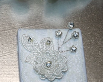 Puede incluir: Un pequeño lienzo blanco con un adorno de encaje blanco y pedrería. El adorno presenta una delicada flor con un centro de pedrería.