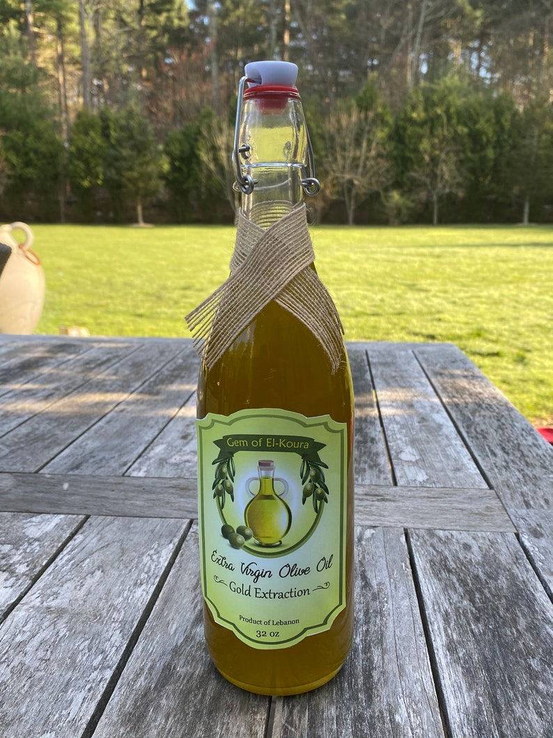 May include: A clear glass bottle of extra virgin olive oil with a gold label that reads "Gem of El-Koura Extra Virgin Olive Oil Gold Extraction Product of Lebanon 32 oz". The bottle is wrapped in burlap and has a red and white flip top.