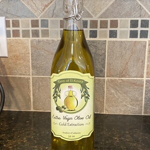 May include: A clear glass bottle of extra virgin olive oil with a green label that reads "Gem of El-Koura Extra Virgin Olive Oil Gold Extraction Product of Lebanon 32 oz".