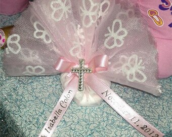 May include: A pink tulle and ribbon favour bag with a white butterfly design. The bag is tied with a pink ribbon and a silver cross charm. The ribbon has the text "Faith Isabella Colon November 11, 2012" printed on it.