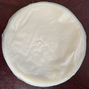 May include: A white round fabric disc with a light blue trim. The fabric is smooth and has a slight sheen.