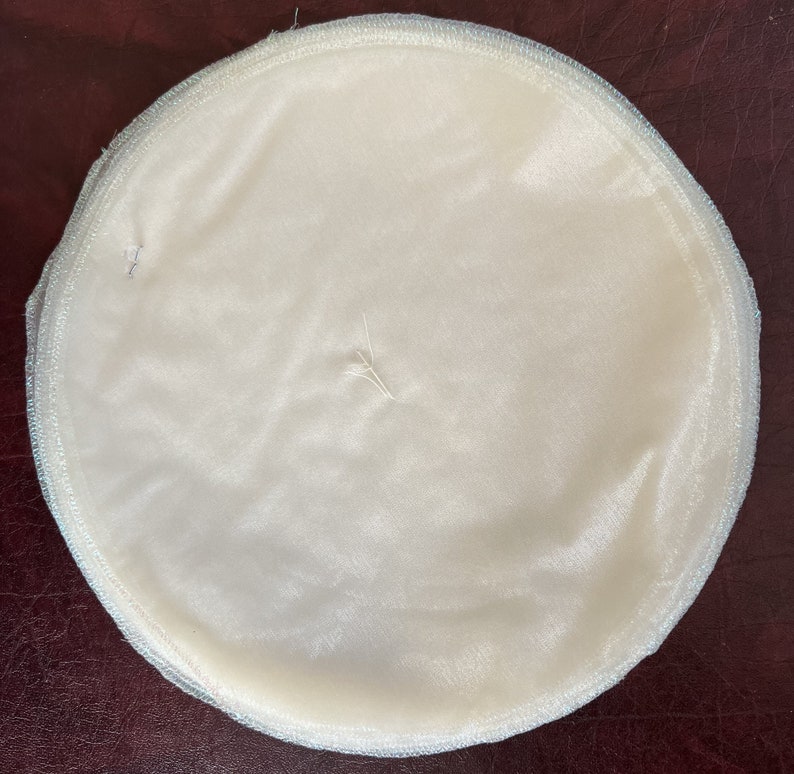 May include: A white round fabric disc with a light blue trim. The fabric is smooth and has a slight sheen.
