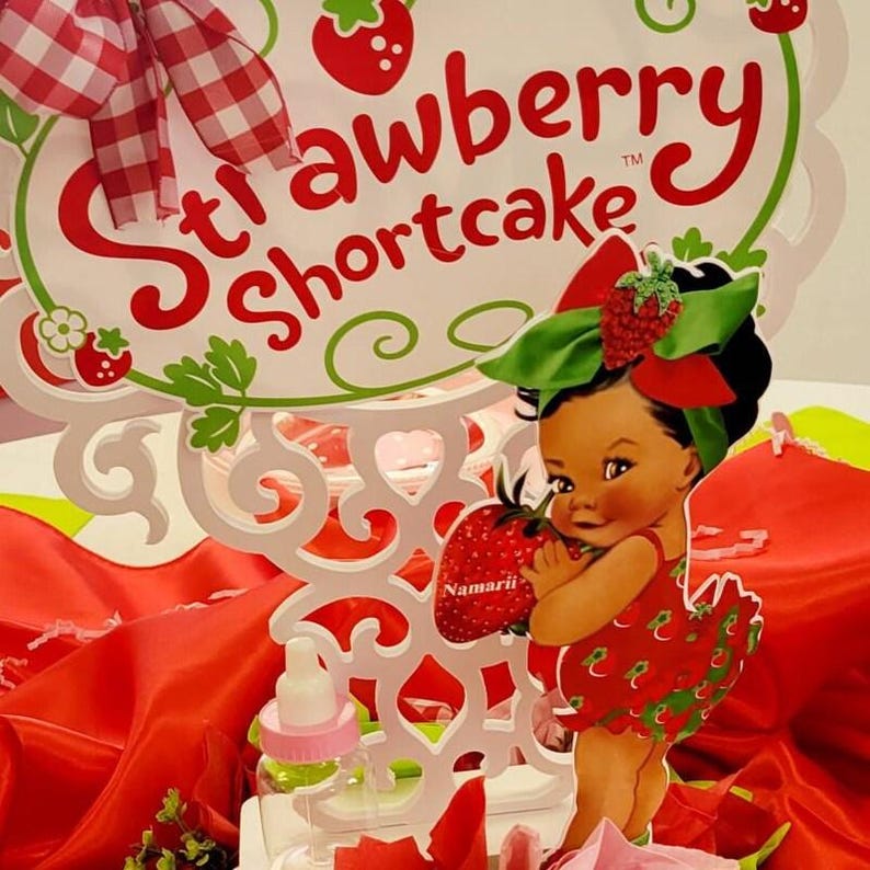 Pu&ograve; includere: Un'insegna decorativa Strawberry Shortcake con cornice bianca intagliata, testo rosso, un fiocco a quadretti rossi e bianchi e l'immagine di un bambino che tiene una grande fragola. Un piccolo biberon in plastica trasparente con tappo rosa &egrave; davanti.