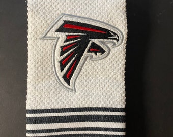 Puede incluir: Toalla de cocina blanca con un logotipo bordado de los Atlanta Falcons en negro y rojo. La toalla tiene un borde de rayas negras y blancas.