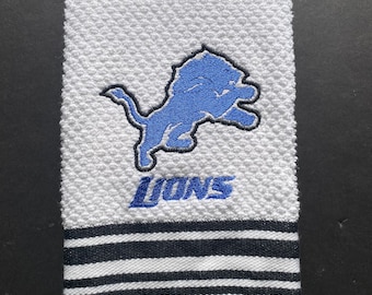 May include: White kitchen towel with a blue embroidered Detroit Lions logo and the word "LIONS" in black. The towel has black and white stripes at the bottom.