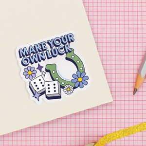 May include: A white notebook with a sticker that says "Make Your Own Luck" with a horseshoe, dice, and flowers. The notebook is on a pink gridded surface with a gold ribbon, a pencil, and a roll of washi tape.