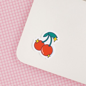 May include: A white laptop computer with a red and green cherry sticker on the lid. The laptop is on a pink grid background.