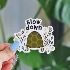 Op de afbeelding: Een witte sticker met een cartoon schildpad en bloemen. De schildpad is groen en heeft een schild met een zeshoekig patroon. De tekst "slow down" staat in het zwart boven de schildpad.