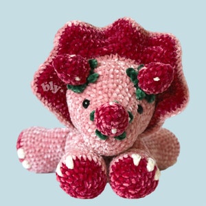 May include: A pink crocheted triceratops stuffed animal with green leaves and red felt flowers. The triceratops has a white star with the word "bly" on its side.