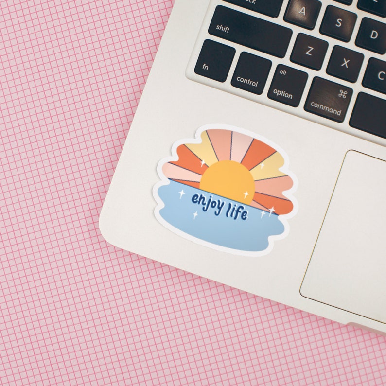May include: A sticker with a sunset over a body of water. The sun is yellow and the sky is orange and pink. The water is blue and there are white stars. The text "enjoy life" is written in blue cursive.