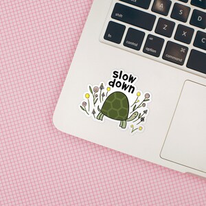 Op de afbeelding: Een witte laptop met een sticker erop. De sticker toont een cartoon schildpad met een groene schild die door een veld met bloemen loopt. De tekst "slow down" staat boven de schildpad.
