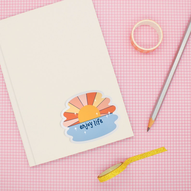 May include: A white notebook with a sticker of a sunset with the text "enjoy life" on a pink grid background. There is a roll of gold washi tape and a pencil on the surface.
