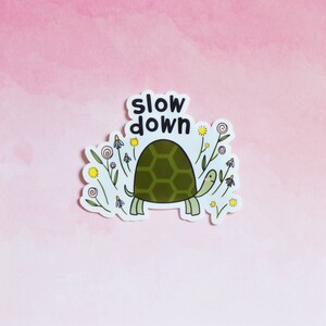 Op de afbeelding: Een sticker met een groene schildpad die door een veld met bloemen loopt. De tekst "slow down" staat boven de schildpad.