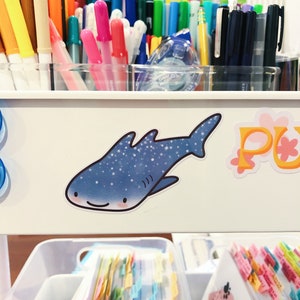 May include: A white plastic organizer with a blue and white whale shark sticker on the front. The organizer has multiple compartments and is filled with colorful markers, pens, and other writing utensils.