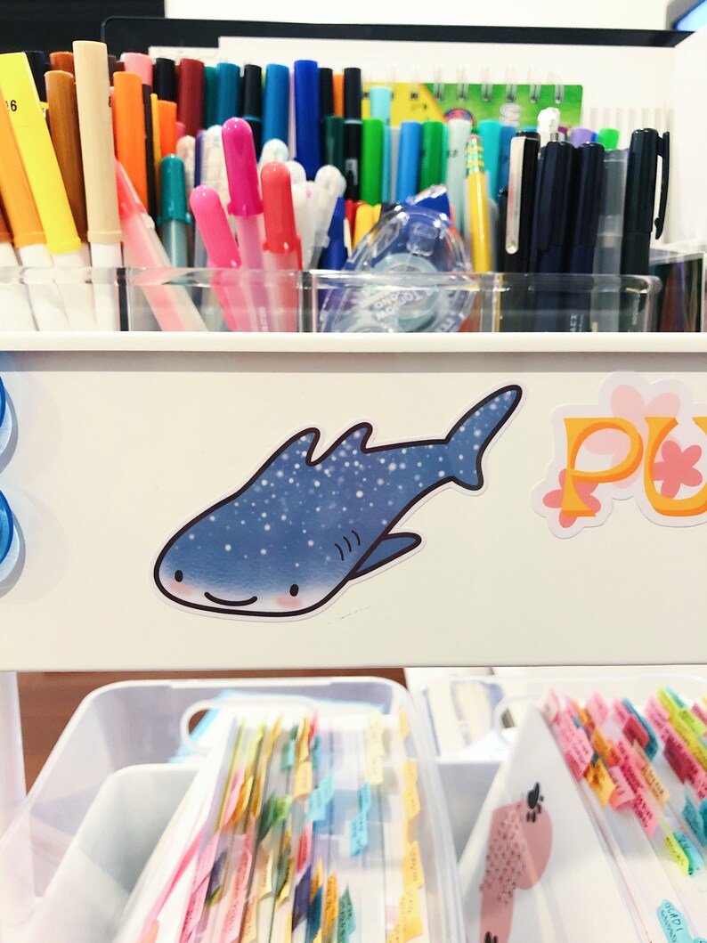 May include: A white plastic organizer with a blue and white whale shark sticker on the front. The organizer has multiple compartments and is filled with colorful markers, pens, and other writing utensils.