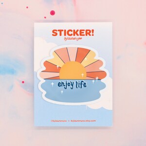 May include: A sticker with a sunset over the ocean and the text "enjoy life". The sticker is in a retro style with a yellow sun, orange and pink stripes, and blue water.