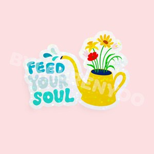 May include: A yellow watering can with a yellow spout and a handle, filled with flowers. The flowers are red, yellow, and orange. The watering can has the text "Feed Your Soul" in blue and white letters.