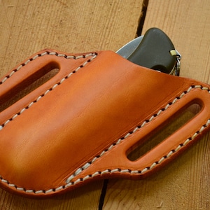 Könnte beinhalten: Eine braune Ledertasche für ein Messer mit weißen Nähten. Die Tasche hat zwei Gürtelschlaufen und ist für ein Klappmesser konzipiert.