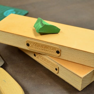 Puede incluir: Dos bloques de cuero apilados para afilar con un compuesto de afilado verde. Los bloques tienen un marco de madera clara y una superficie de cuero marrón claro. La marca "QUERCUR LEATHERCRAFT" es visible en una pequeña etiqueta en cada bloque.