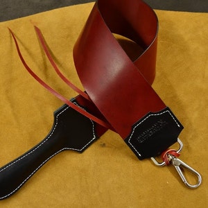 May include: A red leather razor strop with a black leather handle and a silver hook. The strop is folded in half and the handle is on top of the strap. The strap has the text "WIESENBAUM" embossed on it.
