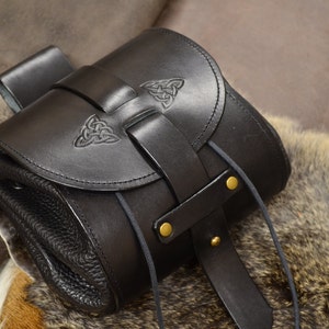 以下が含まれることがあります： フラップ留めとケルト結び目のデザインが施された黒い革製のバッグ。バッグにはストラップと巾着留めがあります。