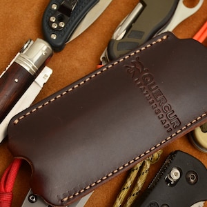 Puede incluir: Una funda de cuchillo de cuero marrón con el texto "Guercuir Leathercraft" grabado en la parte delantera. La funda está cosida con hilo blanco y tiene un cordón rojo atado.