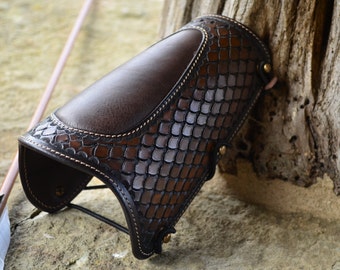 May include: A brown leather arm guard with a textured design. The guard has a strap with a buckle and is secured with a metal snap.