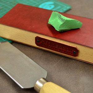 Puede incluir: Una correa de cuero roja con base de madera y una piedra de afilar verde. La correa tiene una etiqueta de cuero con el texto "TOUERCUR" en letras doradas.