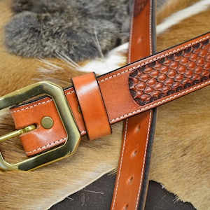 May include: A brown leather belt with a brass buckle and a tooled design. The belt is on a brown and white fur surface.