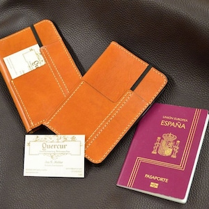 Può includere: Due porta passaporto in pelle arancione aperti con un elastico nero, un biglietto da visita e un passaporto spagnolo. I porta passaporto hanno cuciture bianche e una tasca. Il biglietto da visita riporta il nome "Quercu".