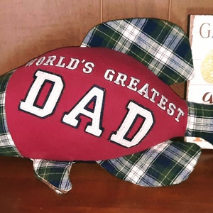May include: A fish-shaped decorative pillow with a red center and plaid green and blue accents. The center features the text "WORLD'S GREATEST DAD" in white lettering. A button serves as an eye.