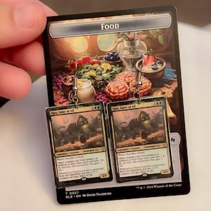 May include: Silver-toned earrings featuring a rectangular design with a fantasy card theme. The earrings display the text "Ygra, Eater of All" and other card details. The background shows a card with the word "FOOD" at the top.