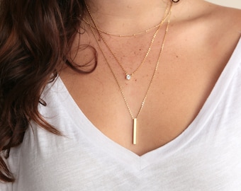 May include: A layered gold necklace with a small diamond pendant and a long, thin bar pendant.