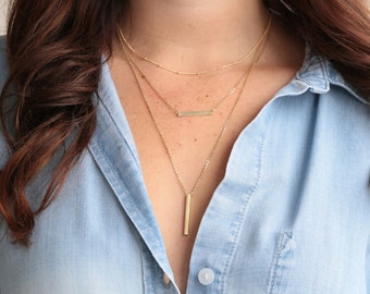 Puede incluir: Un conjunto de collares dorados en capas. La cadena superior tiene pequeñas cuentas, la del medio una barra horizontal y la inferior un colgante de barra largo y vertical. Se muestra sobre una camisa vaquera azul claro.