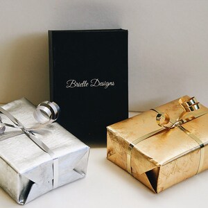 May include: Three gift boxes wrapped in different colors: silver, gold, and black. The black box has the text "Brielle Designs" printed on it.